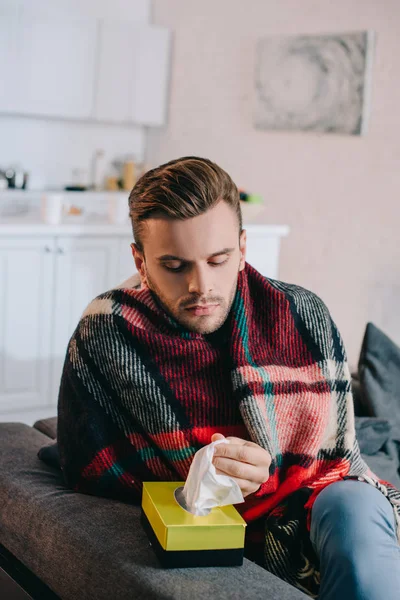 Sick young man taking paper napkins from box while sitting on couch — Stock Photo