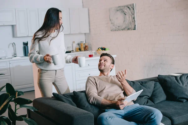 Beautiful young couple with cups of cocoa and tablet relaxing together at home — Stock Photo