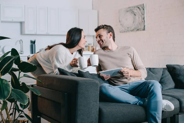 Happy young couple with cups of cocoa and tablet relaxing together at home — Stock Photo