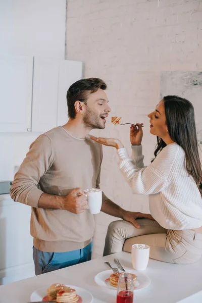 Happy young woman feeding her boyfriend with pancakes for breakfast — Stock Photo