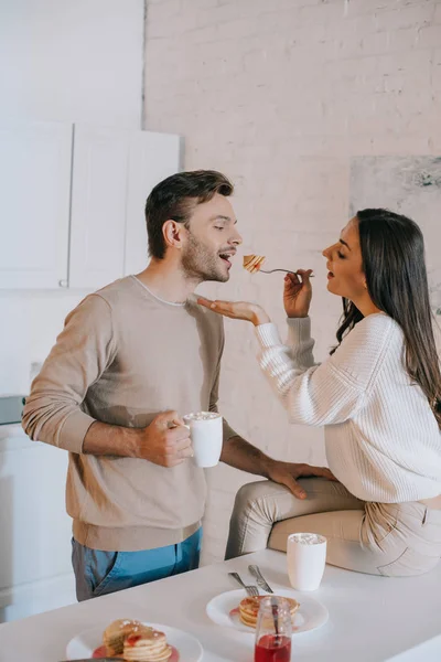 Heureuse jeune femme nourrir son petit ami avec des crêpes pour le petit déjeuner — Photo de stock