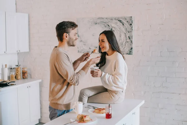 Happy young man feeding his girlfriend with pancakes for breakfast — Stock Photo