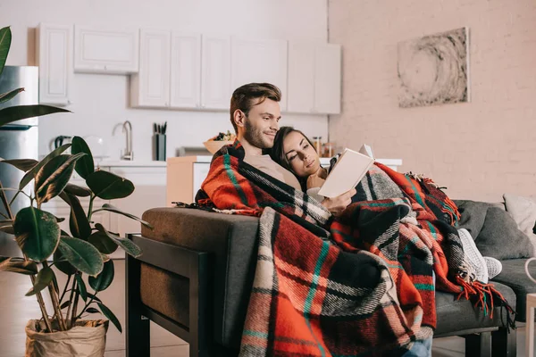 Sonriente joven pareja leyendo libro juntos en sofá bajo cuadros en casa - foto de stock