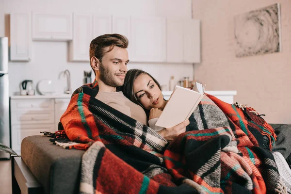 Feliz joven pareja leyendo libro juntos en sofá bajo cuadros en casa - foto de stock