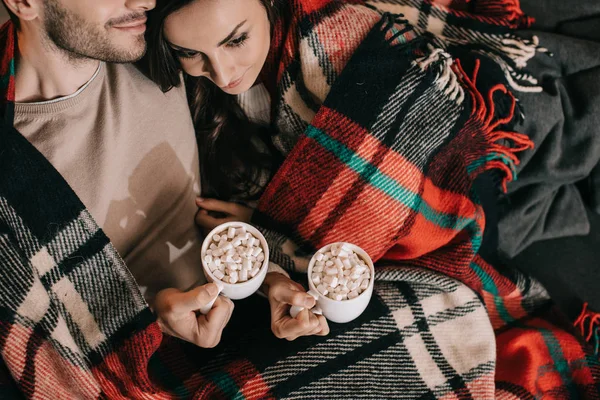 Vista superior de pareja joven con tazas de cacao con malvavisco relajándose en el sofá bajo cuadros en casa - foto de stock