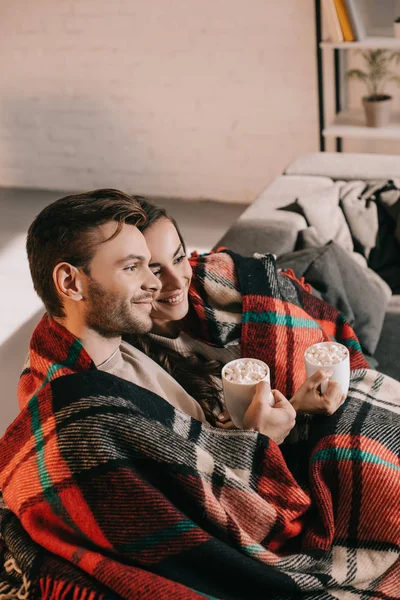 Smiling young couple with cups of cocoa with marshmallow relaxing on couch under plaid at home — Stock Photo