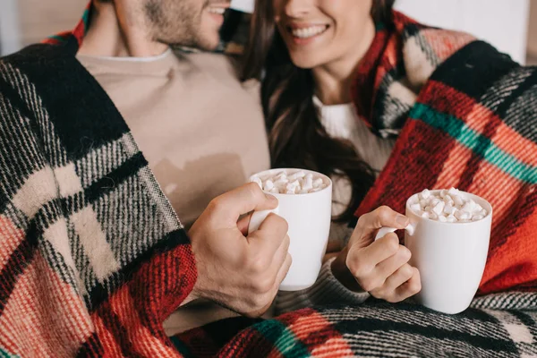 Cropped shot of smiling young couple with cups of cocoa with marshmallow relaxing on couch under plaid at home — Stock Photo