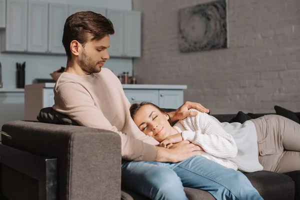 Beau jeune homme assis sur le canapé tandis que sa petite amie dort sur lui à la maison — Photo de stock