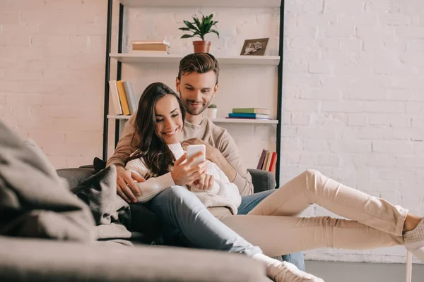 Sorrindo jovem casal usando smartphone juntos no sofá em casa — Fotografia de Stock