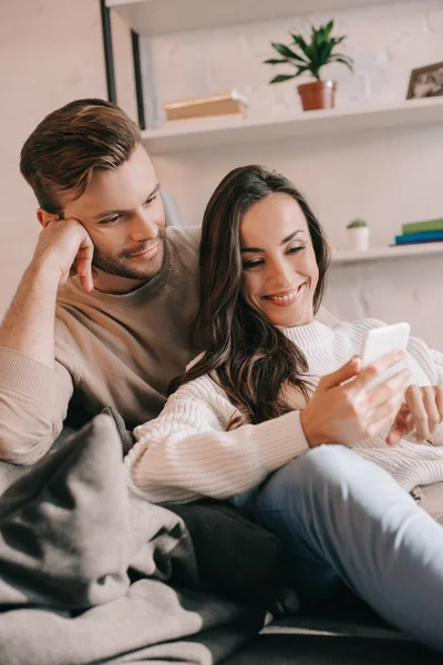 Sorrindo jovem casal usando smartphone juntos no sofá em casa — Fotografia de Stock