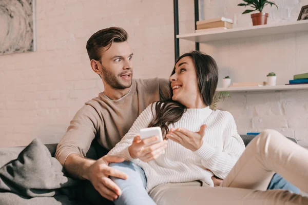 Überraschtes junges Paar nutzt Smartphone gemeinsam auf der heimischen Couch — Stockfoto