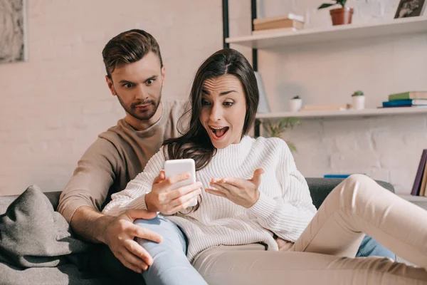 Expressivo jovem casal usando smartphone juntos no sofá em casa — Fotografia de Stock