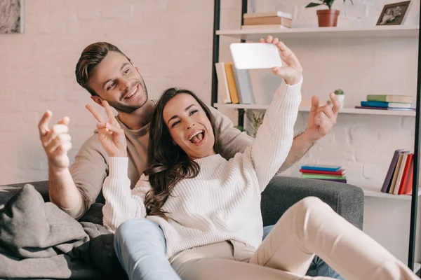 Feliz joven pareja tomando selfie con teléfono inteligente y haciendo gesto de paz en el sofá en casa - foto de stock