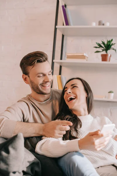 Смеющаяся молодая пара с помощью смартфона вместе на диване дома — стоковое фото