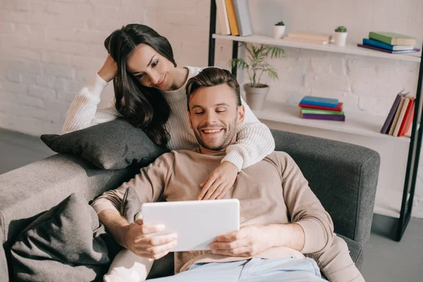 Sorrindo jovem casal usando tablet juntos no sofá em casa — Fotografia de Stock