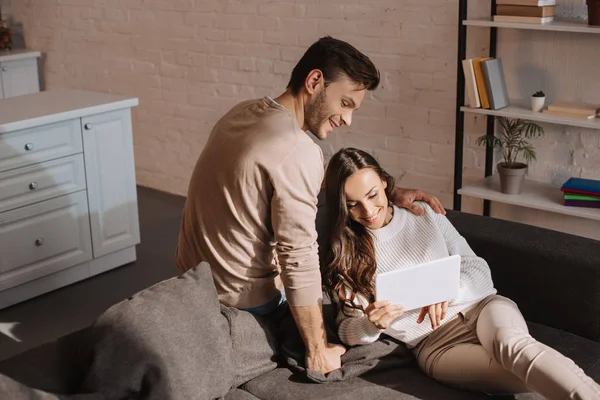 Sourire jeune couple en utilisant la tablette ensemble sur le canapé à la maison — Photo de stock