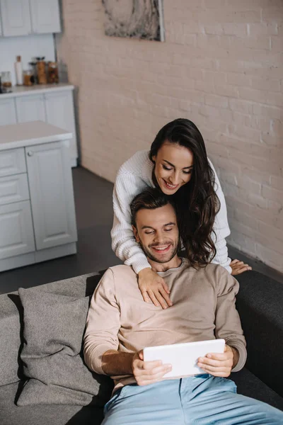 Vue grand angle de jeune femme heureuse embrassant son petit ami par derrière alors qu'il utilise une tablette à la maison — Photo de stock