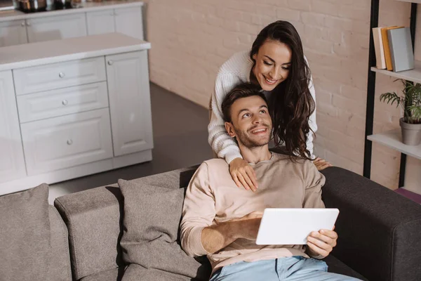 Vue grand angle de jeune femme attrayante embrassant son petit ami par derrière alors qu'il utilise une tablette à la maison — Photo de stock