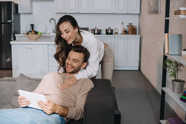 Beautiful young woman embracing her boyfriend from behind while he using tablet at home — Stock Photo