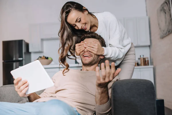 Beautiful young woman covering eyes of boyfriend while he using tablet at home — Stock Photo