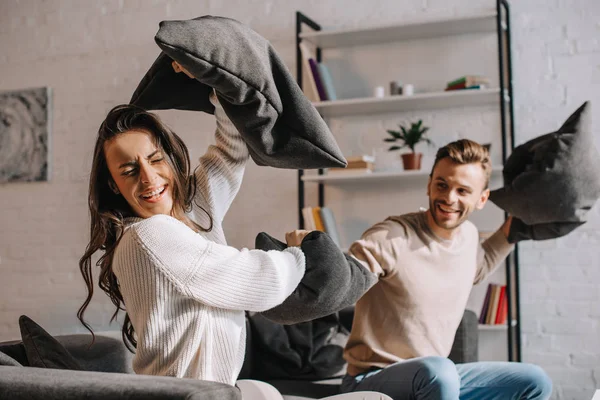 Feliz jovem casal lutando com travesseiros no sofá em casa — Fotografia de Stock