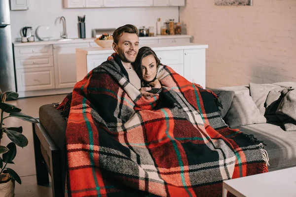 Feliz joven pareja relajarse en el sofá y ver la televisión juntos mientras cubre con cuadros - foto de stock