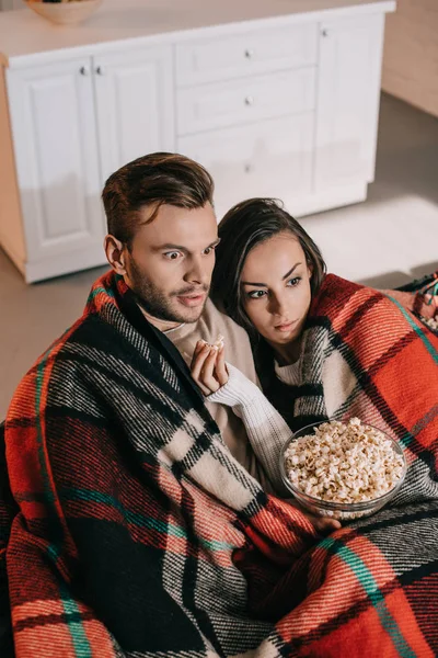 Vista de ángulo alto de la joven pareja impactada viendo la película con palomitas de maíz en el sofá y cubriendo con cuadros - foto de stock