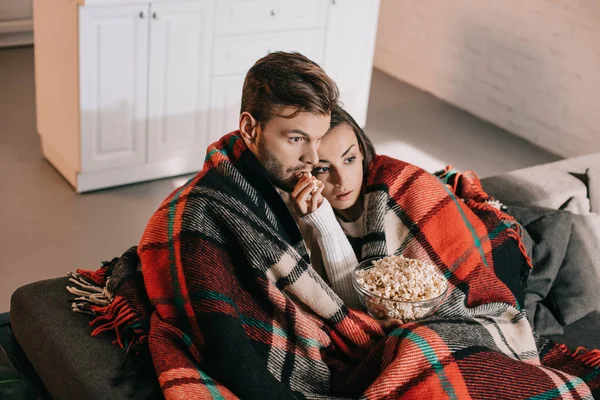 Vista de ángulo alto de la joven pareja emocional viendo la película con palomitas de maíz en el sofá y cubierta con cuadros - foto de stock
