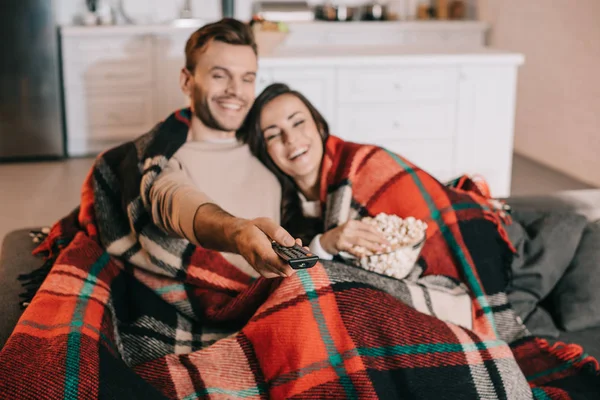 Feliz joven pareja viendo la televisión con palomitas de maíz en el sofá y cubriendo con cuadros - foto de stock