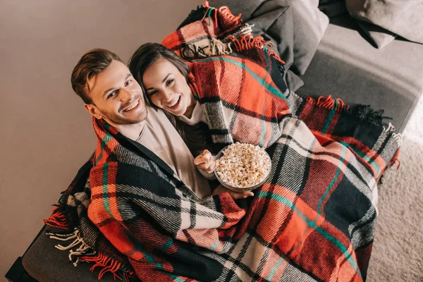 Vista de ángulo alto de pareja joven relajándose en el sofá con un tazón de palomitas de maíz y cubriendo con cuadros - foto de stock