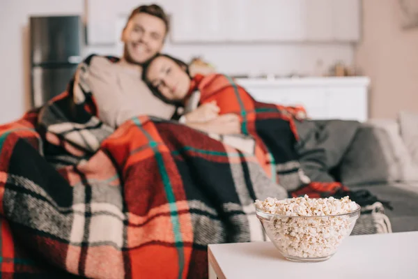Hermosa pareja joven relajándose en el sofá y cubriendo con cuadros con tazón de palomitas de maíz en primer plano - foto de stock
