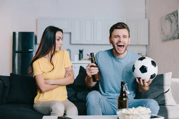 Junge Frau sieht ihren Mann kritisch an, während er Fußballspiel schaut und zu Hause jubelt — Stockfoto