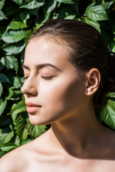 Young beautiful woman with closed eyes against sunshine with green leaves at background — Stock Photo