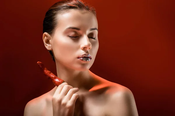 Mujer atractiva joven con labios brillantes, ojos cerrados y chile rojo sobre fondo borgoña - foto de stock