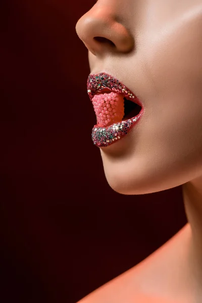 Крупним планом молода жінка з блискучими губами тримає рожеві цукерки в роті на бордовому фоні — стокове фото