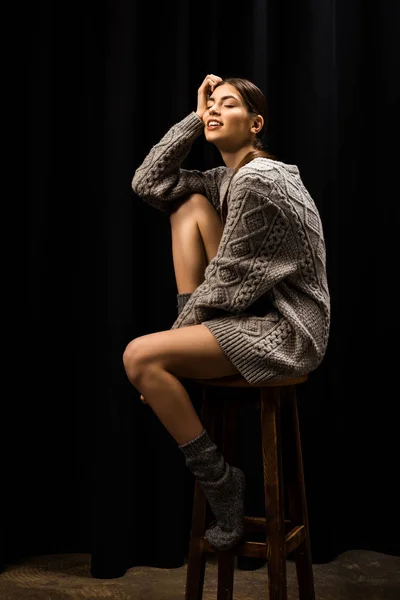 Young smiling woman in woolen grey sweater and socks sitting on wooden bar stool on black background — Stock Photo