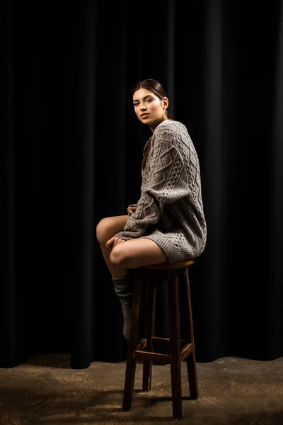 Young pretty woman in woolen grey sweater and socks on bar stool on black background — Stock Photo
