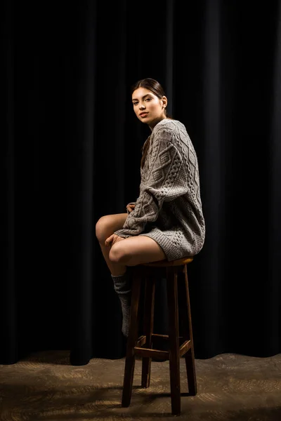 Jeune jolie femme en laine gris pull et chaussettes sur tabouret de bar sur fond noir — Photo de stock