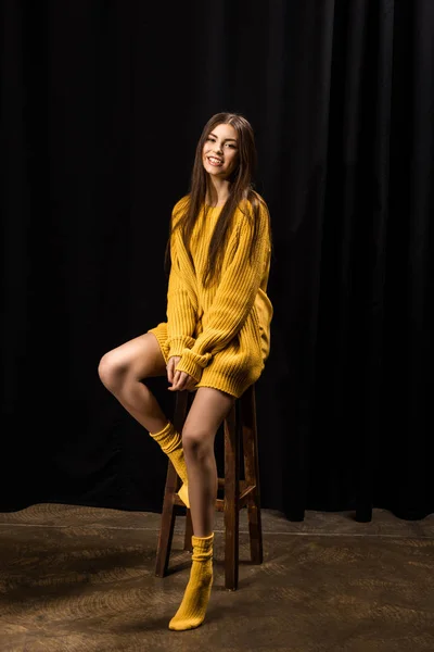 Femme souriante en laine jaune pull assis sur tabouret de bar sur fond noir — Photo de stock