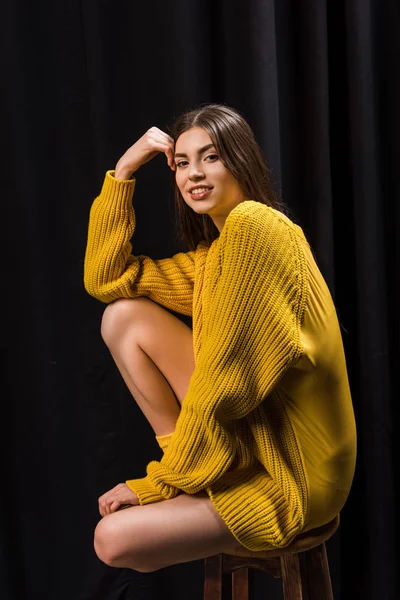 Side view of smiling woman in yellow woolen sweater on wooden bar stool on black backdrop — Stock Photo