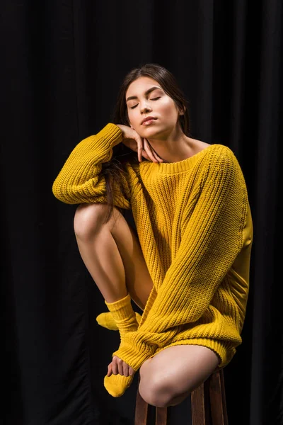 Woman in yellow woolen sweater with eyes closed sitting on bar stool on black backdrop — Stock Photo