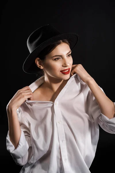 Elegante modelo joven con labios rojos en camisa blanca y sombrero negro posando aislado en negro - foto de stock