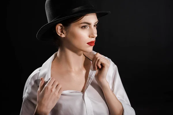Portrait of beautiful pensive woman in white shirt and black hat posing isolated on black — Stock Photo