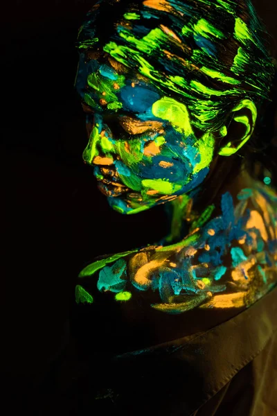 Vista lateral de la mujer pintada con pinturas ultravioletas de colores sobre fondo negro - foto de stock
