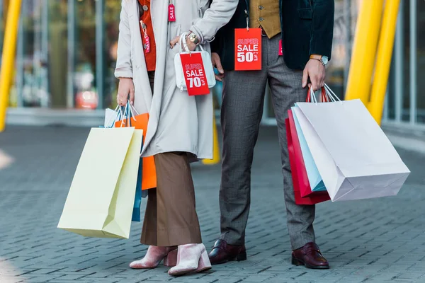 Vue recadrée du couple élégant élégant avec des sacs à provisions et des étiquettes de vente posant près du centre commercial — Photo de stock