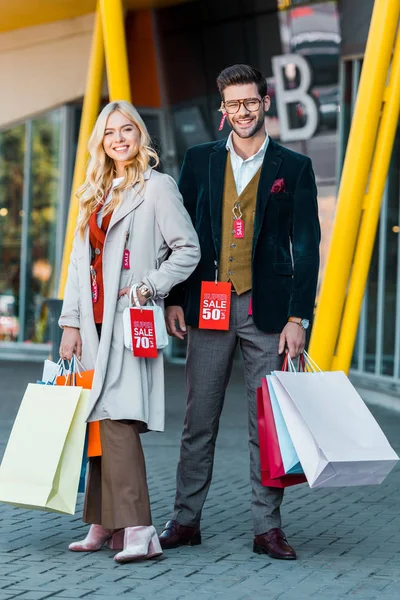 Heureux couple élégant avec des sacs à provisions et des étiquettes de vente posant près du centre commercial — Photo de stock