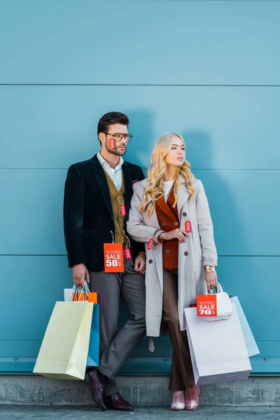 Coppia elegante che tiene borse della spesa con tag di vendita e posa al muro blu — Foto stock