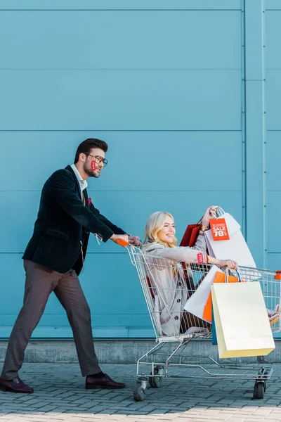 Belo casal elegante com sacos de compras e etiquetas de venda se divertindo com carrinho de compras na parede azul — Fotografia de Stock