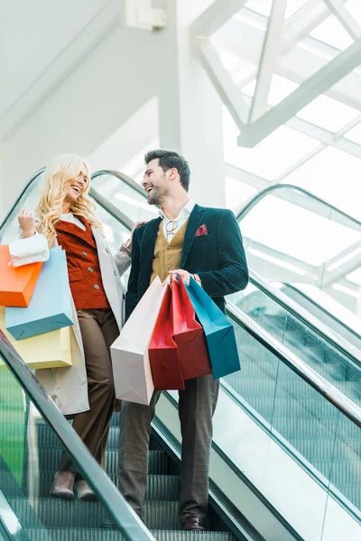 Happy stylish couple with shopping bags standing on escalator — Stock Photo