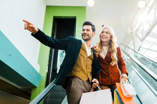 Smiling stylish man showing something to girlfriend with shopping bags standing on escalator — Stock Photo